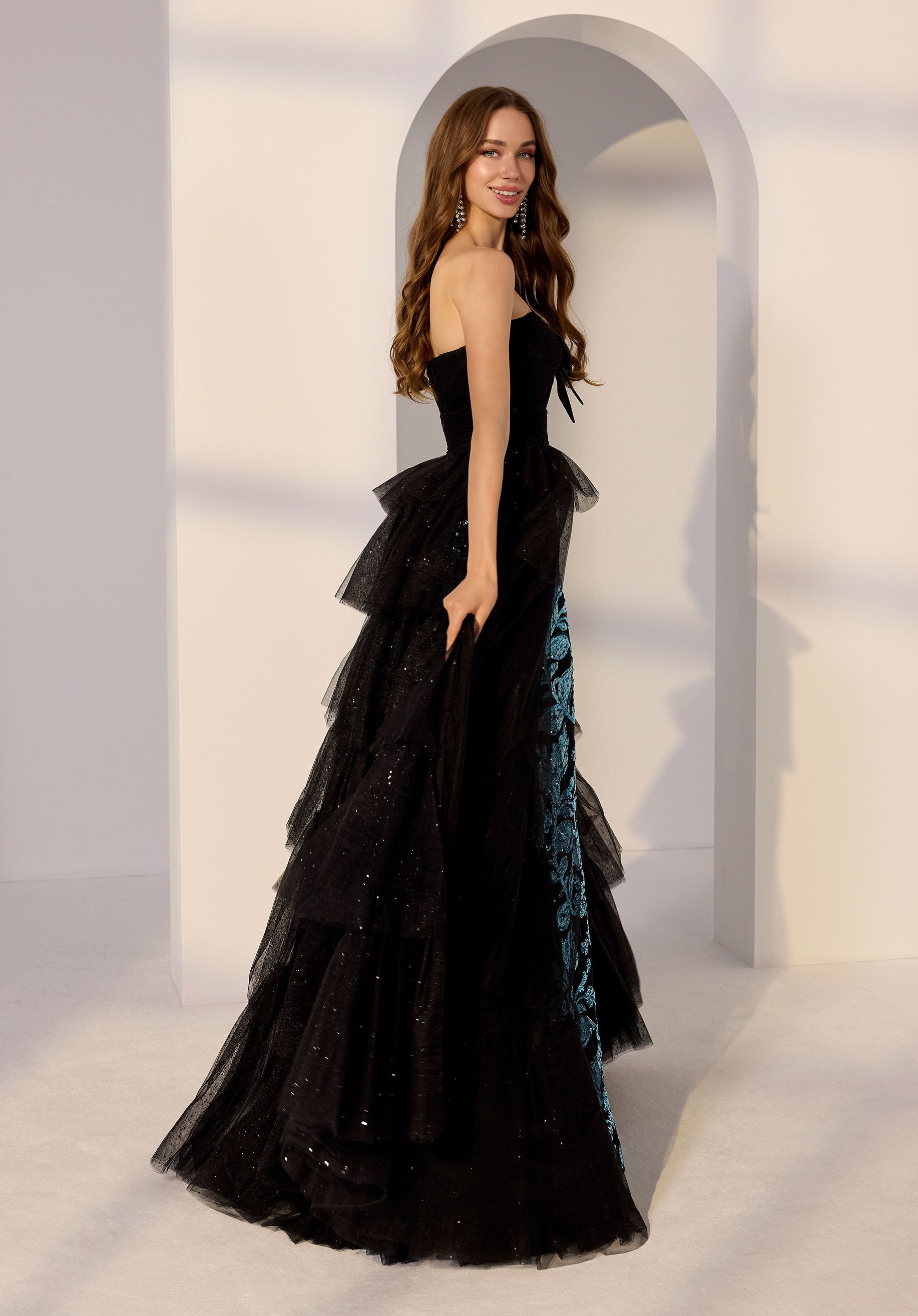 A woman wearing the Morilee 49275 long black strapless prom dress with blue details, layered tulle, and a detachable overskirt stands before a white arched doorway, glancing over her shoulder.